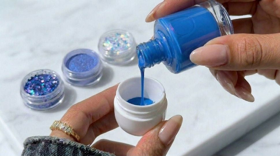A close-up of a person pouring bright blue nail polish into a small white jar, with three small clear containers of blue glitter and shimmer visible on a light marble surface.