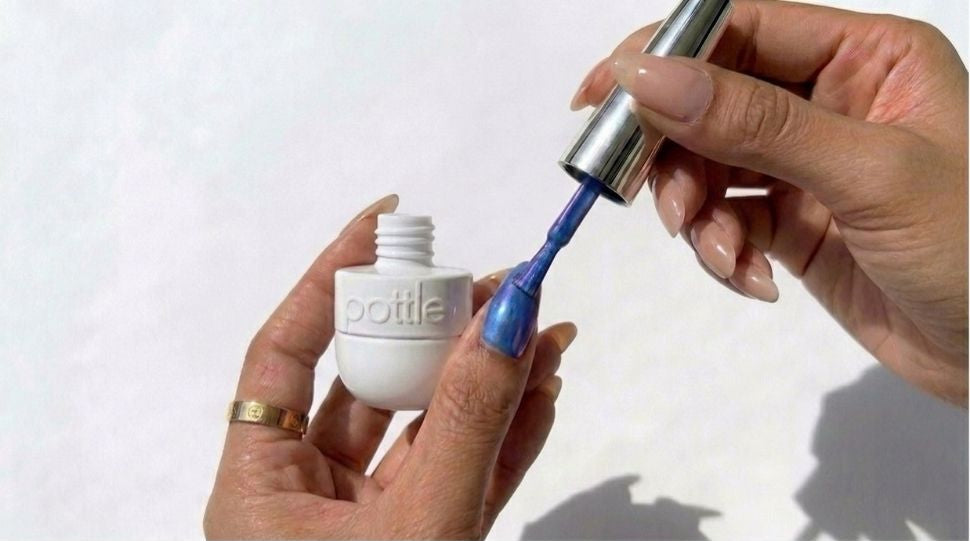 A close-up of a person's hands holding a white bottle labeled 'pottle' while applying bright blue nail polish with a silver-handled brush.